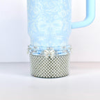 A light blue tumbler with a floral and bow pattern. The bottom section is covered in silver-colored faux pearls. A light blue handle is attached to the side. The bottom of the tumbler has a beaded design with a silver bow.