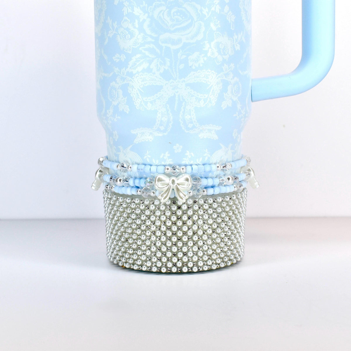A light blue tumbler with a floral and bow pattern. The bottom section is covered in silver-colored faux pearls. A light blue handle is attached to the side. The bottom of the tumbler has a beaded design with a silver bow.