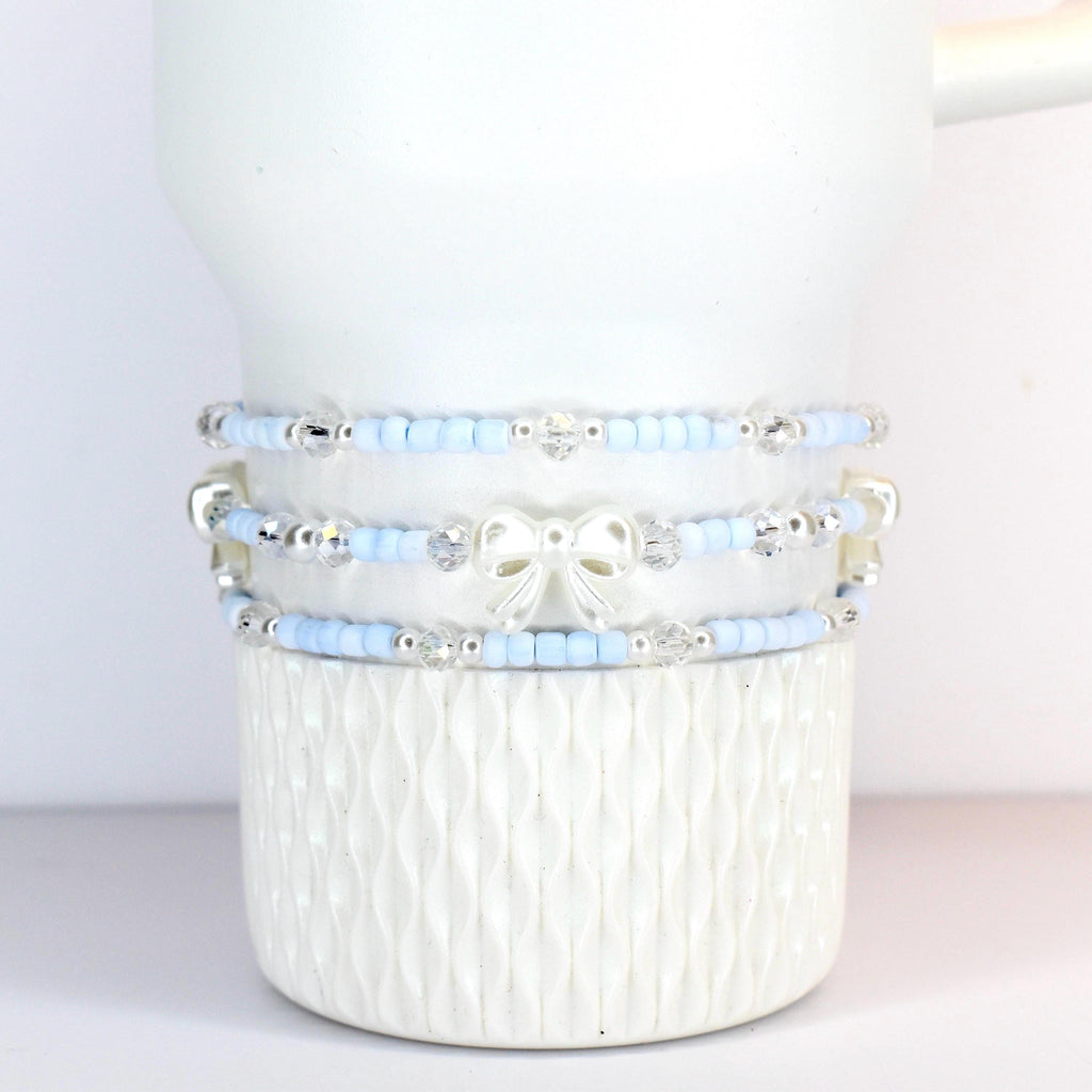 Three beaded bracelets in light blue and silver, adorned with clear crystals and a white bow. The bracelets are stacked on a white, textured ceramic mug. The beads appear to be small and delicate.