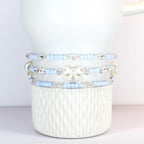 Three beaded bracelets in light blue and silver, adorned with clear crystals and a white bow. The bracelets are stacked on a white, textured ceramic mug. The beads appear to be small and delicate.