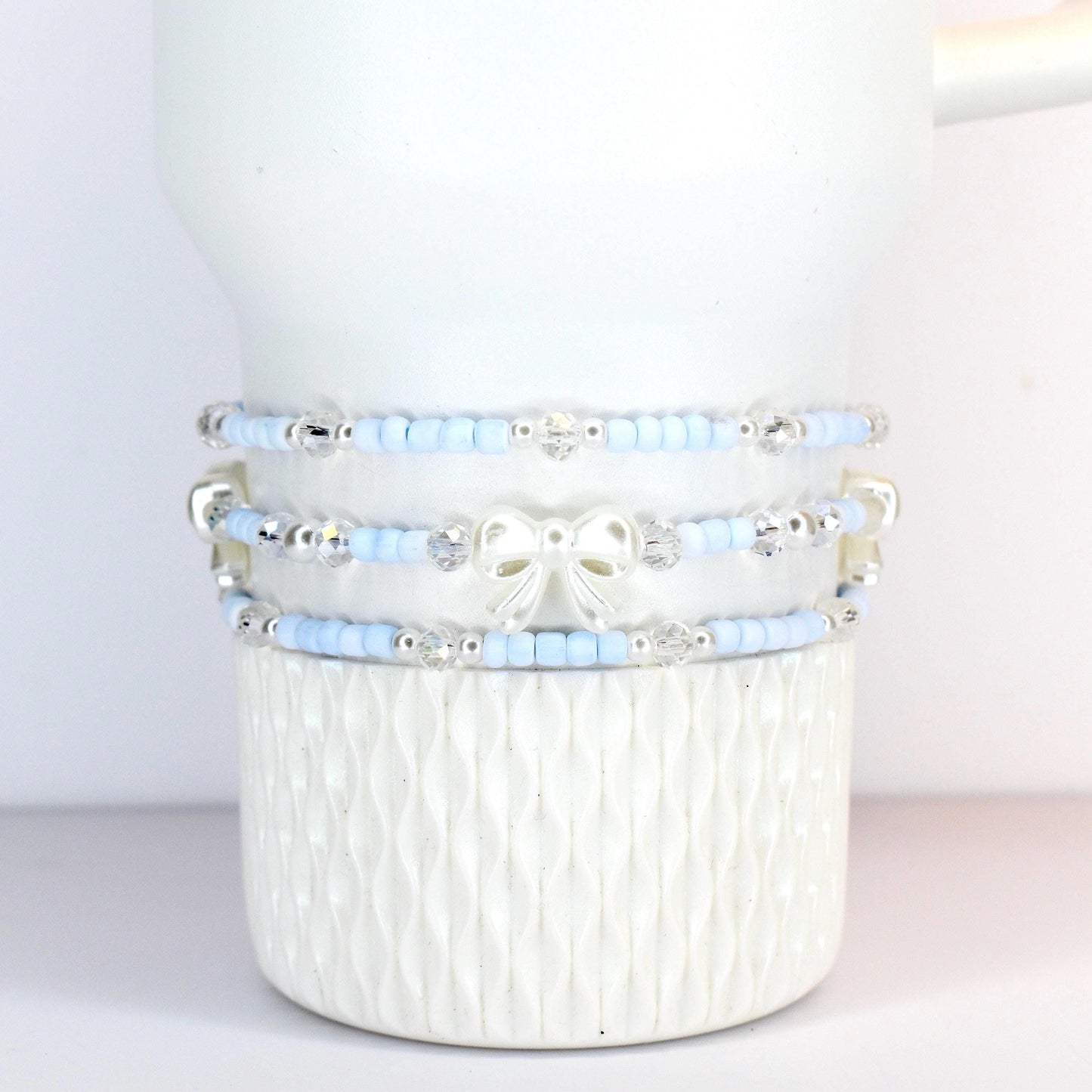 Three beaded bracelets in light blue and silver, adorned with clear crystals and a white bow. The bracelets are stacked on a white, textured ceramic mug. The beads appear to be small and delicate.