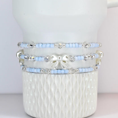Three-piece bracelet set. Each bracelet features light blue beads, clear crystal accents, and silver-toned details. The middle bracelet has a white bow charm. The bracelets are stacked on a white, textured surface.
