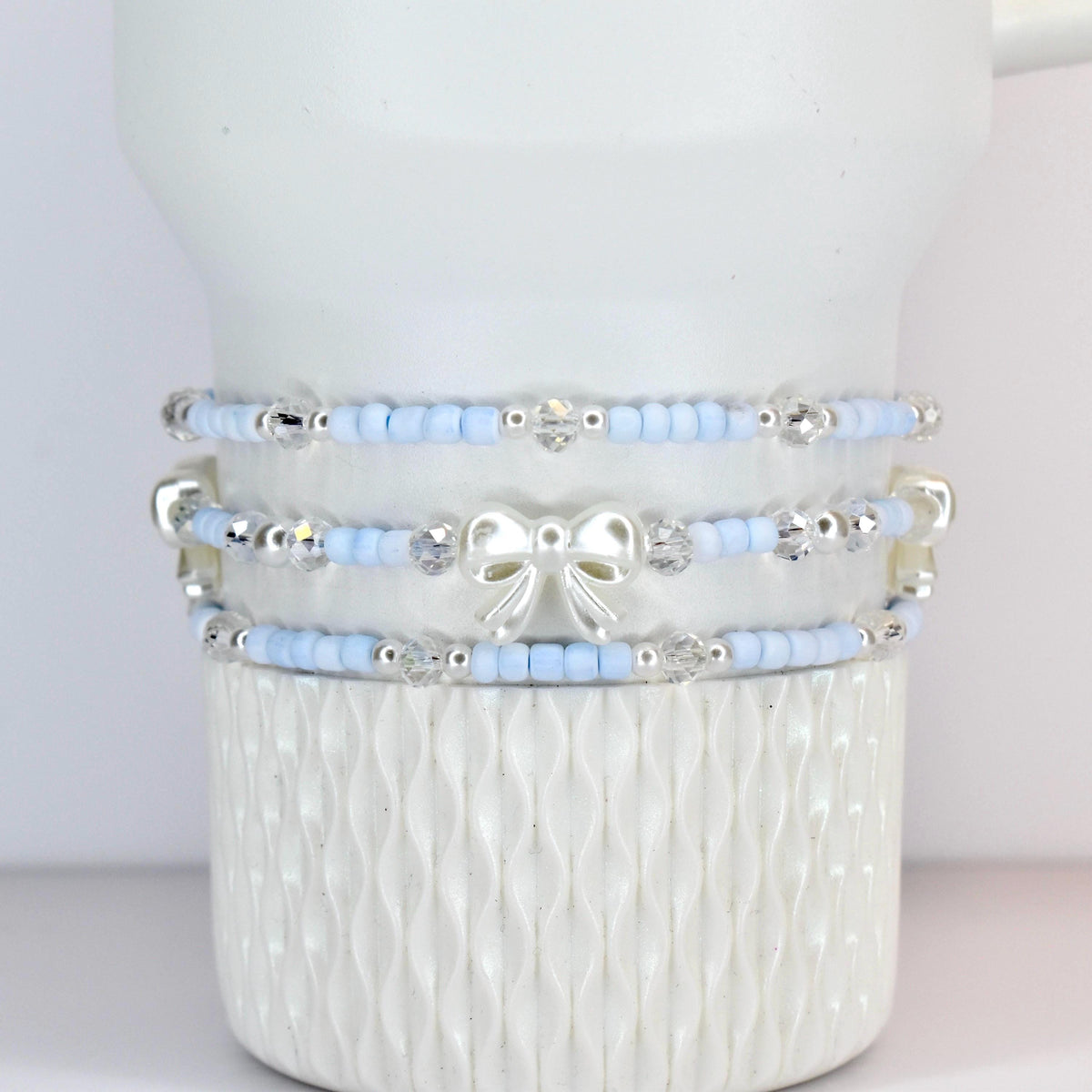 Three-piece bracelet set. Each bracelet features light blue beads, clear crystal accents, and silver-toned details. The middle bracelet has a white bow charm. The bracelets are stacked on a white, textured surface.