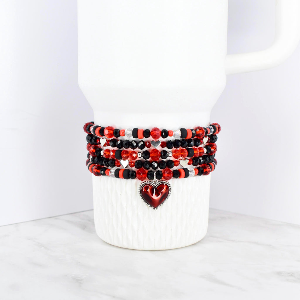 A stack of beaded bracelets in red, black, and silver, wrapped around a white tumbler. The bracelets feature a variety of beads and a heart-shaped charm. The tumbler is white with a handle.