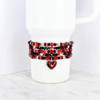 A stack of beaded bracelets in red, black, and silver, wrapped around a white tumbler. The bracelets feature a variety of beads and a heart-shaped charm. The tumbler is white with a handle.