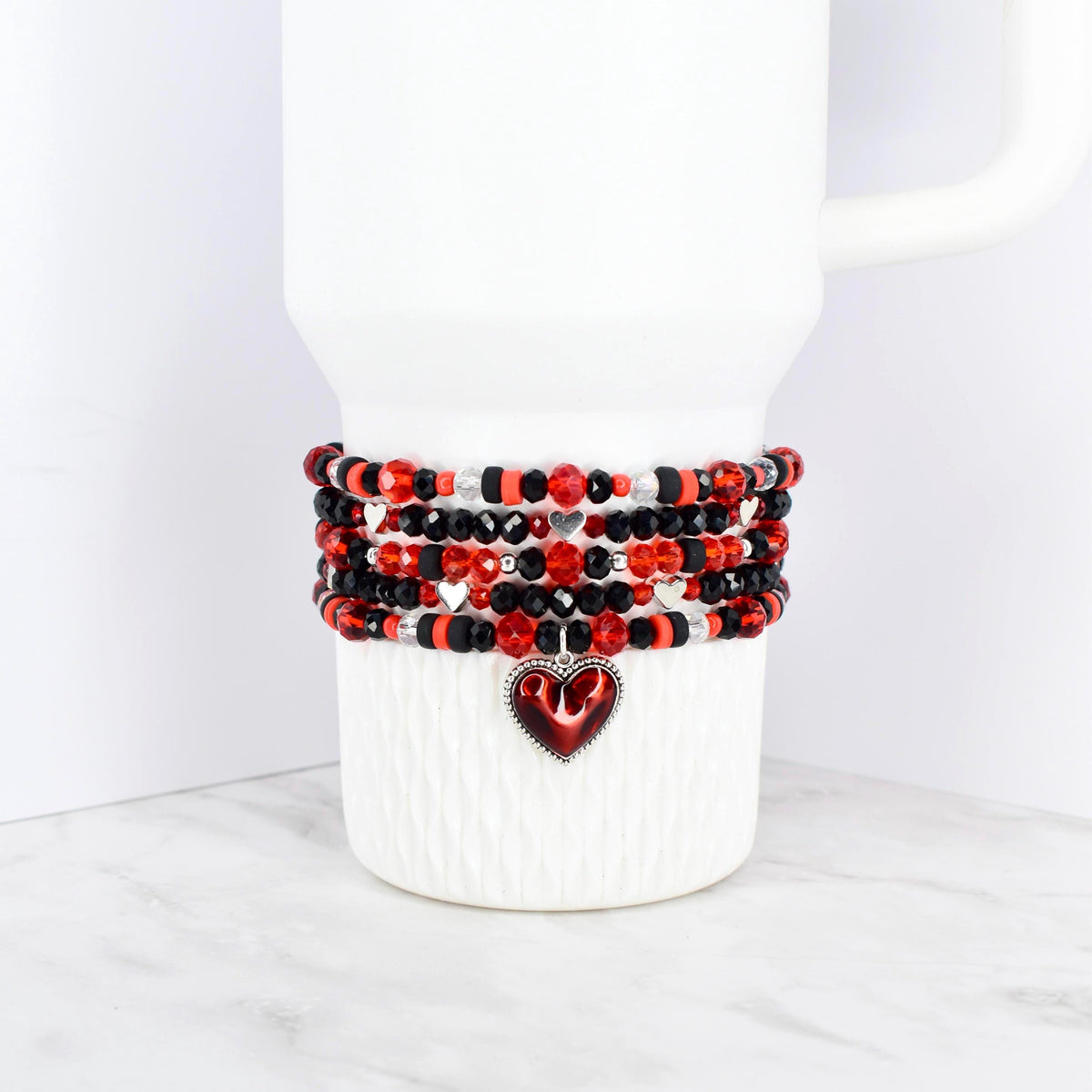 A stack of beaded bracelets in red, black, and silver, wrapped around a white tumbler. The bracelets feature a variety of beads and a heart-shaped charm. The tumbler is white with a handle.