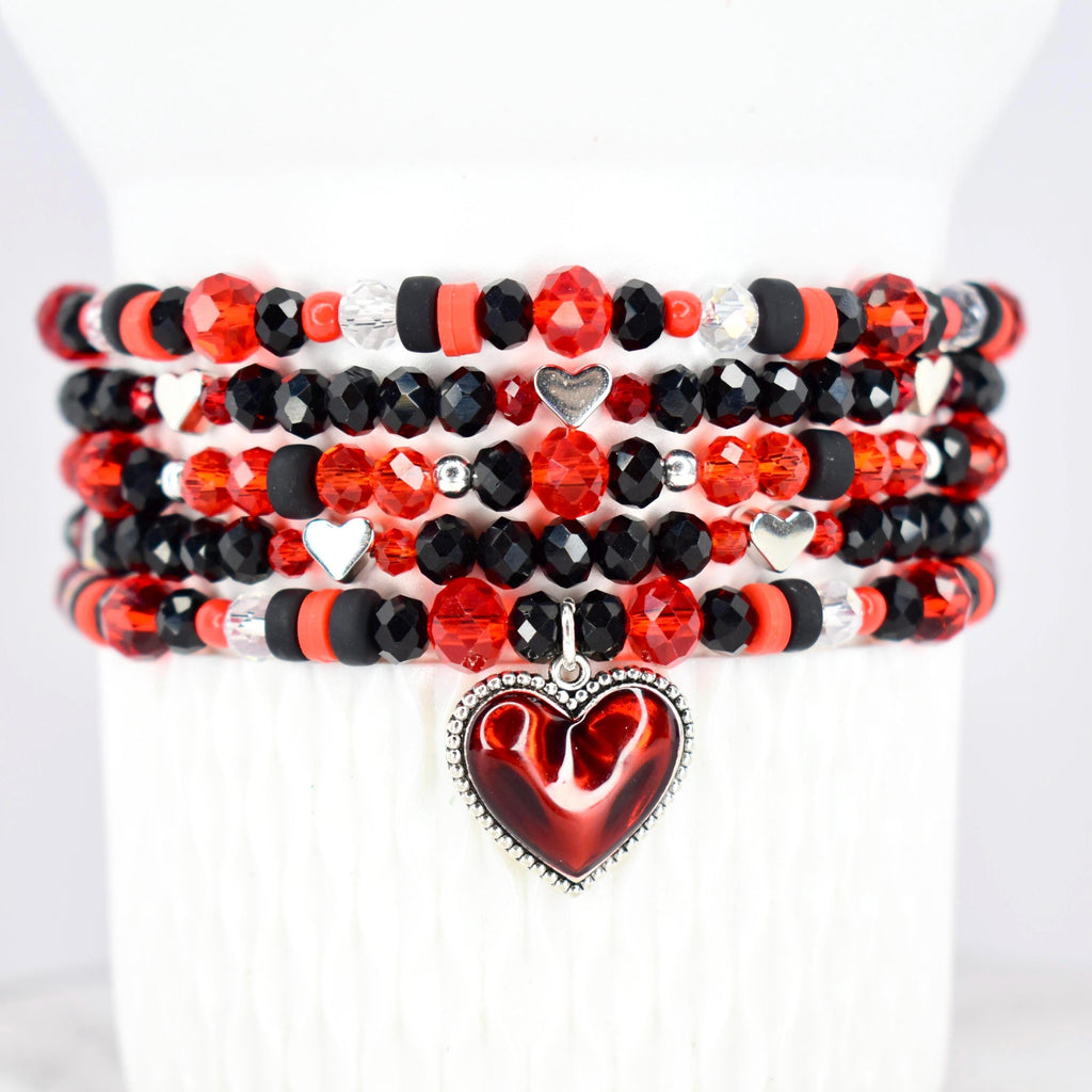Stack of beaded bracelets in red, black, and clear crystal beads. Each bracelet features a mix of faceted and round beads, with silver heart charms. A red heart pendant with a silver border hangs from the bottom bracelet.
