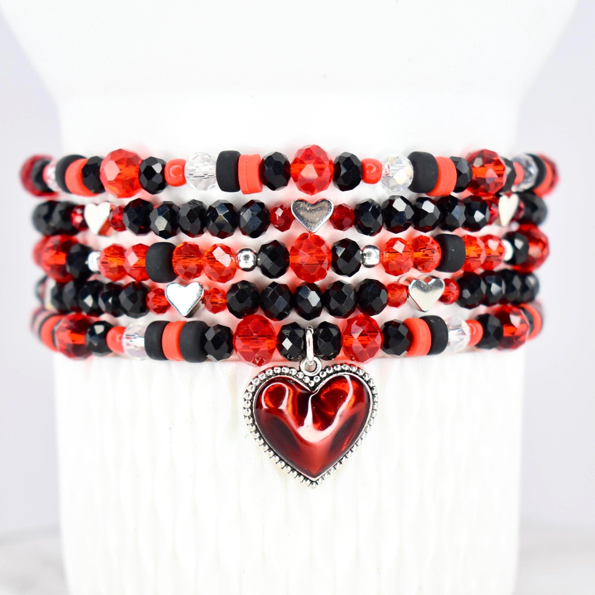 Stack of beaded bracelets in red, black, and clear crystal beads. Each bracelet features a mix of faceted and round beads, with silver heart charms. A red heart pendant with a silver border hangs from the bottom bracelet.