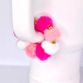 A colorful tumbler handle bracelet featuring fluffy pom-poms in shades of pink, white, and gold accents. The pom-poms are attached to a gold-colored metal band. The bracelet is held against a white background.