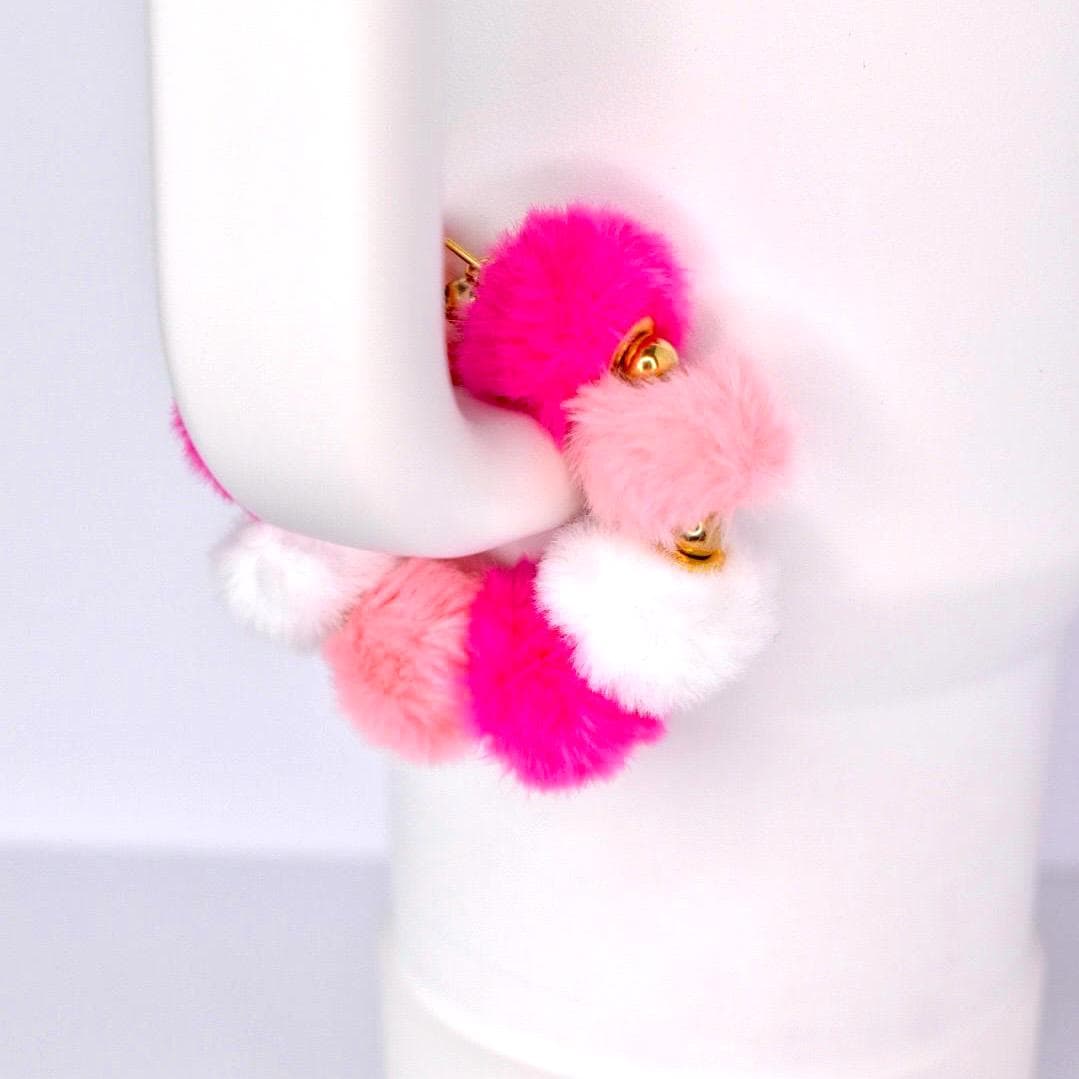 A colorful tumbler handle bracelet featuring fluffy pom-poms in shades of pink, white, and gold accents. The pom-poms are attached to a gold-colored metal band. The bracelet is held against a white background.
