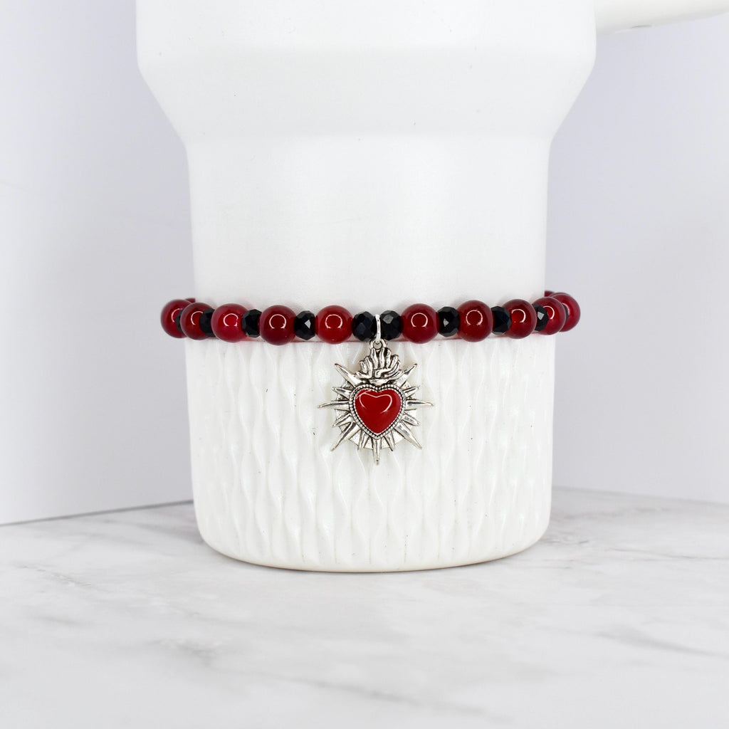 A beaded bracelet featuring red and black beads. The bracelet has a silver-toned charm with a red heart at the center, surrounded by a sunburst design. The bracelet is displayed on a white, textured surface.