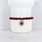 A beaded bracelet featuring red and black beads. The bracelet has a silver-toned charm with a red heart at the center, surrounded by a sunburst design. The bracelet is displayed on a white, textured surface.