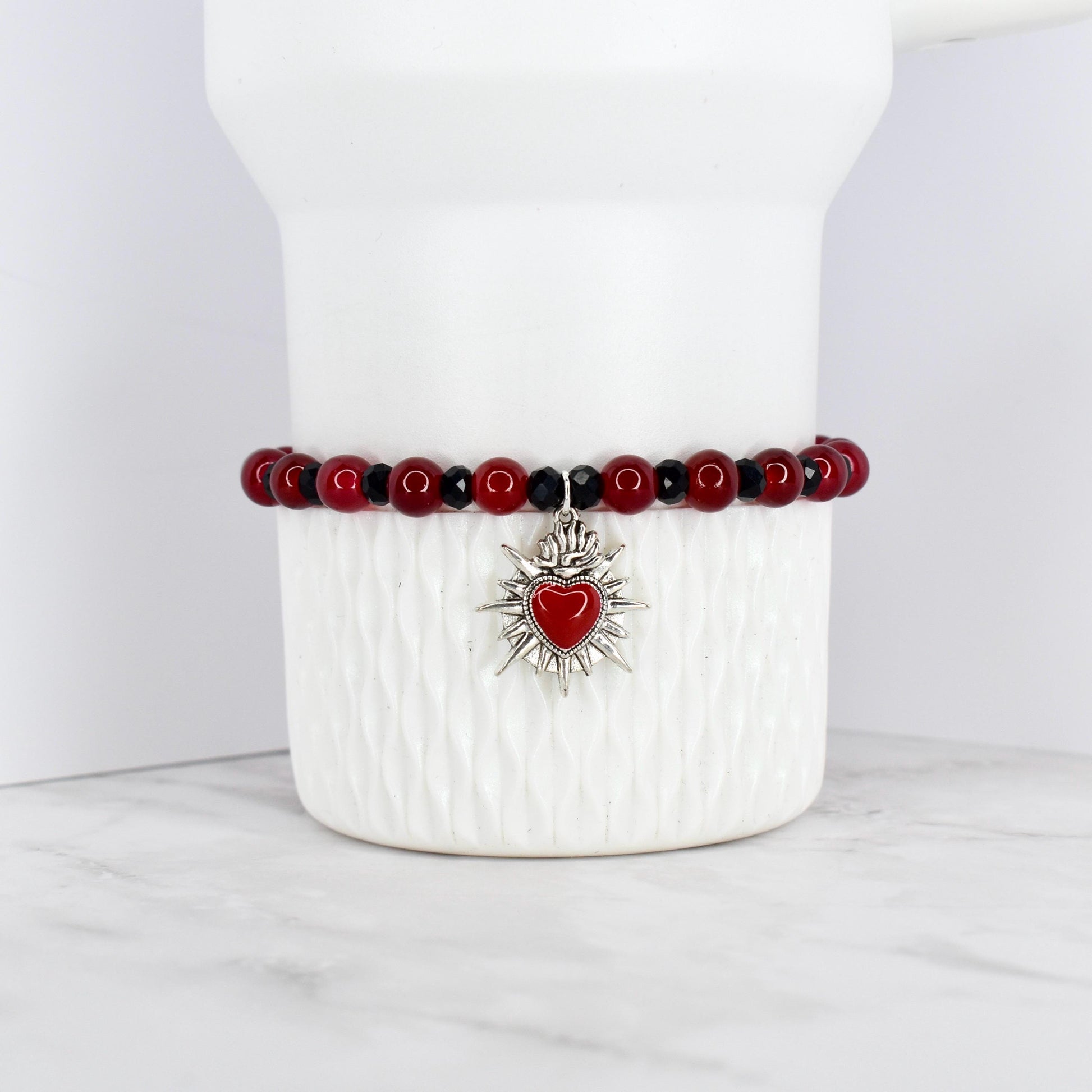 A beaded bracelet featuring red and black beads. The bracelet has a silver-toned charm with a red heart at the center, surrounded by a sunburst design. The bracelet is displayed on a white, textured surface.