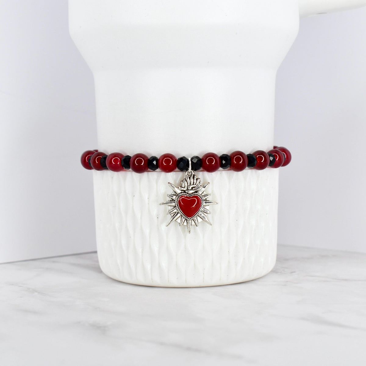 A beaded bracelet featuring red and black beads. The bracelet has a silver-toned charm with a red heart at the center, surrounded by a sunburst design. The bracelet is displayed on a white, textured surface.