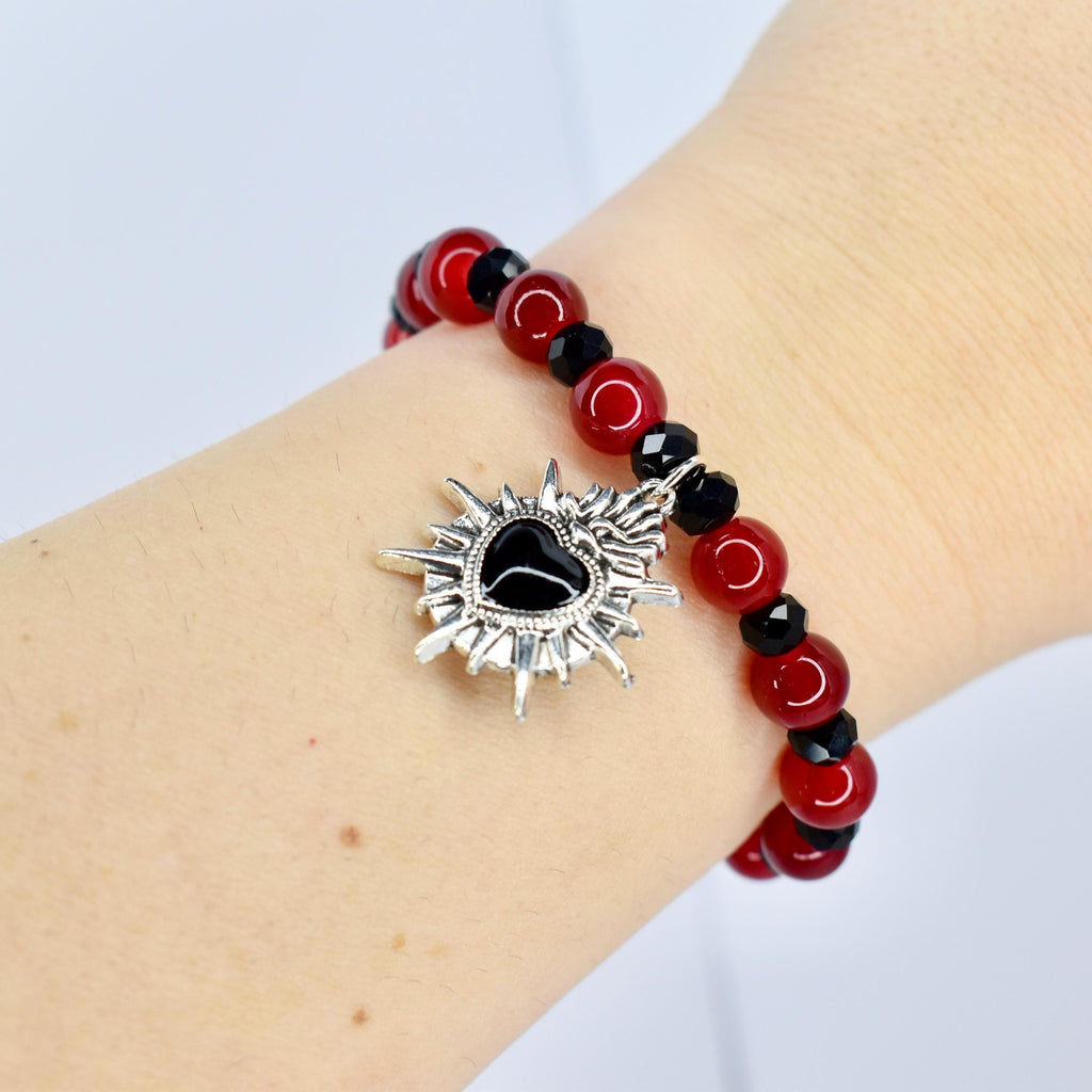 A red beaded bracelet with black accents and a silver charm. The charm is a sunburst design with a black heart in the center. The bracelet is worn on a wrist.