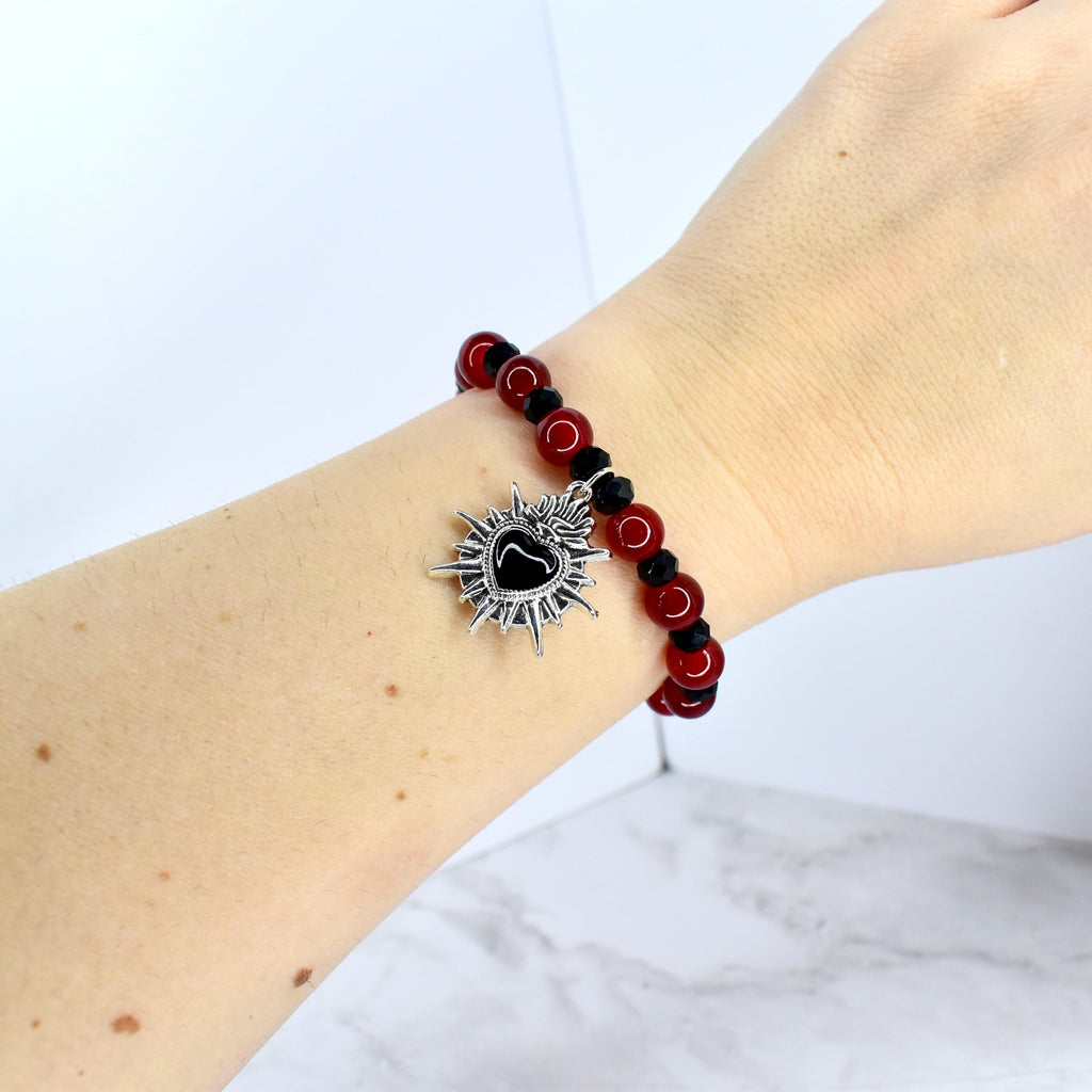A beaded bracelet featuring red and black beads. A silver-tone charm with a black heart center and sunburst design hangs from the bracelet. The bracelet is worn on a wrist.