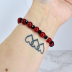 A beaded bracelet featuring alternating red and black beads. The red beads are round and polished, while the black beads are faceted. The bracelet is worn on a wrist with a tattoo of three connected hearts.
