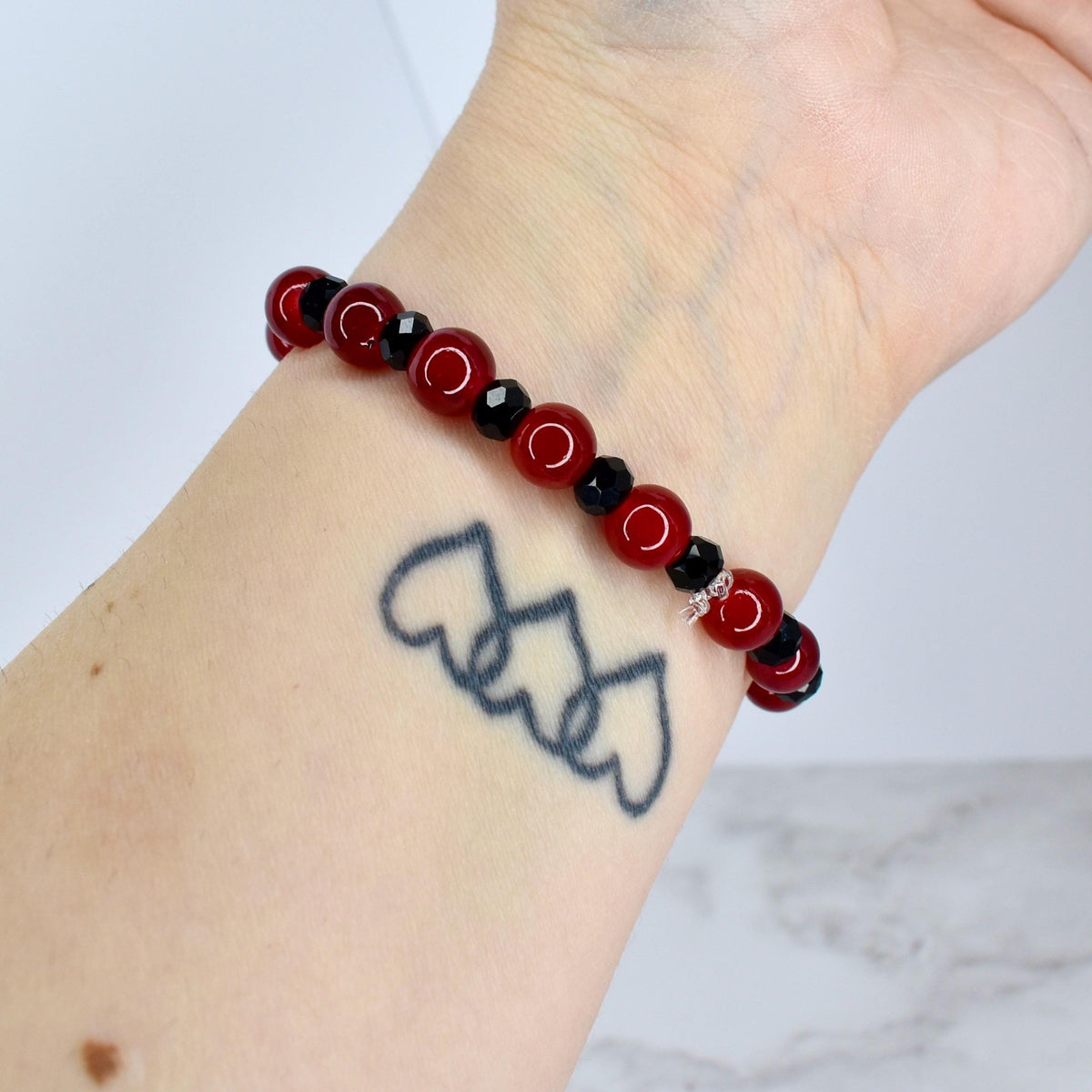 A beaded bracelet featuring alternating red and black beads. The red beads are round and polished, while the black beads are faceted. The bracelet is worn on a wrist with a tattoo of three connected hearts.