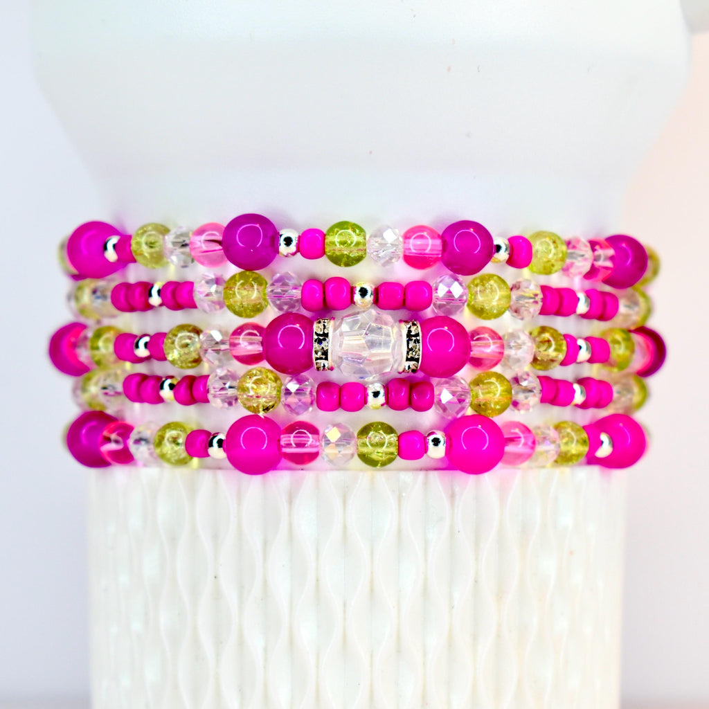 Stack of beaded bracelets in shades of pink, green, and clear. The bracelets feature a variety of bead sizes and textures, including large, round pink beads, smaller pink beads, and clear and green beads. Silver-colored spacers are also present.