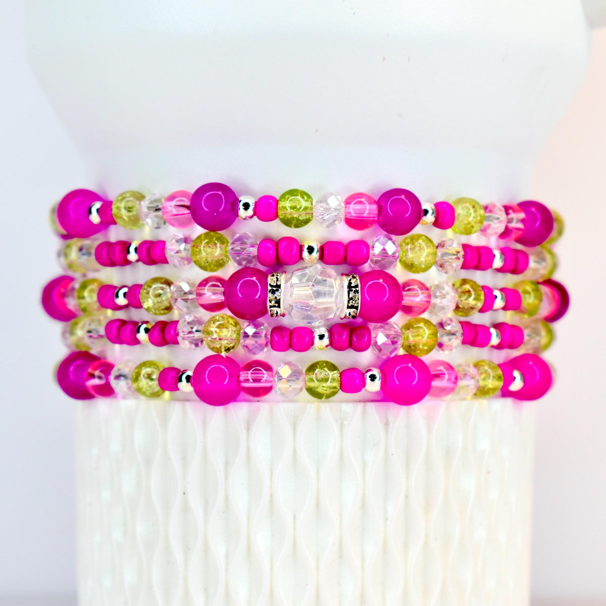 Stack of beaded bracelets in shades of pink, green, and clear. The bracelets feature a variety of bead sizes and textures, including large, round pink beads, smaller pink beads, and clear and green beads. Silver-colored spacers are also present.