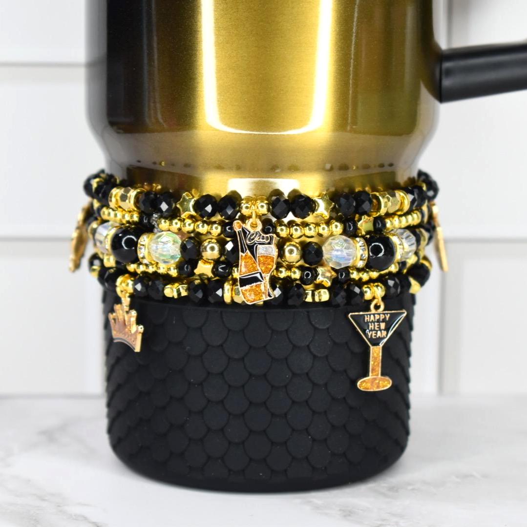 a black honey Stanley tumbler with a set of five black and gold bracelets around the base. the bracelets have new years themed charms from two of the bracelets.