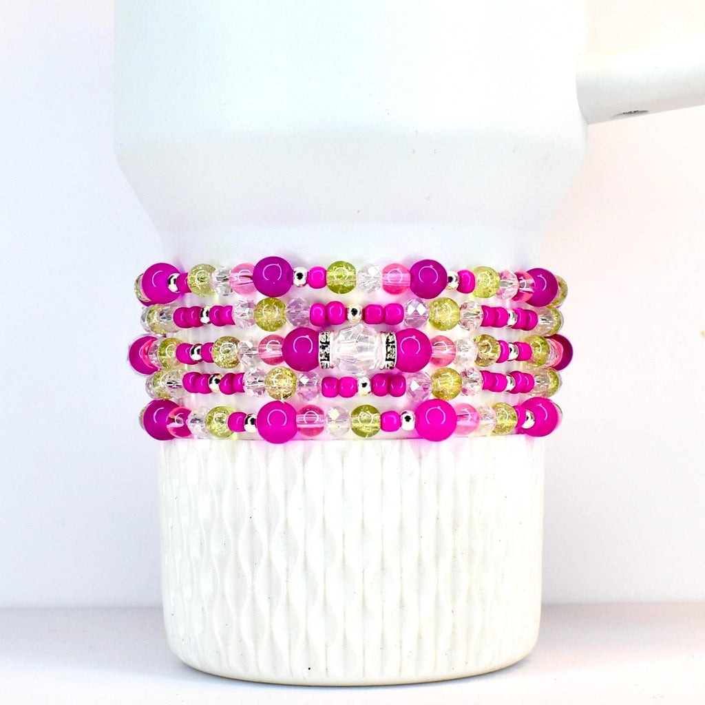 Stack of beaded bracelets in shades of pink, green, and clear. The bracelets are made with various sizes of beads, including large pink orbs, small silver beads, and clear faceted beads. The bracelets are stacked on a white tumbler.