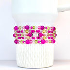 Stack of beaded bracelets in shades of pink, green, and clear. The bracelets are made with various sizes of beads, including large pink orbs, small silver beads, and clear faceted beads. The bracelets are stacked on a white tumbler.