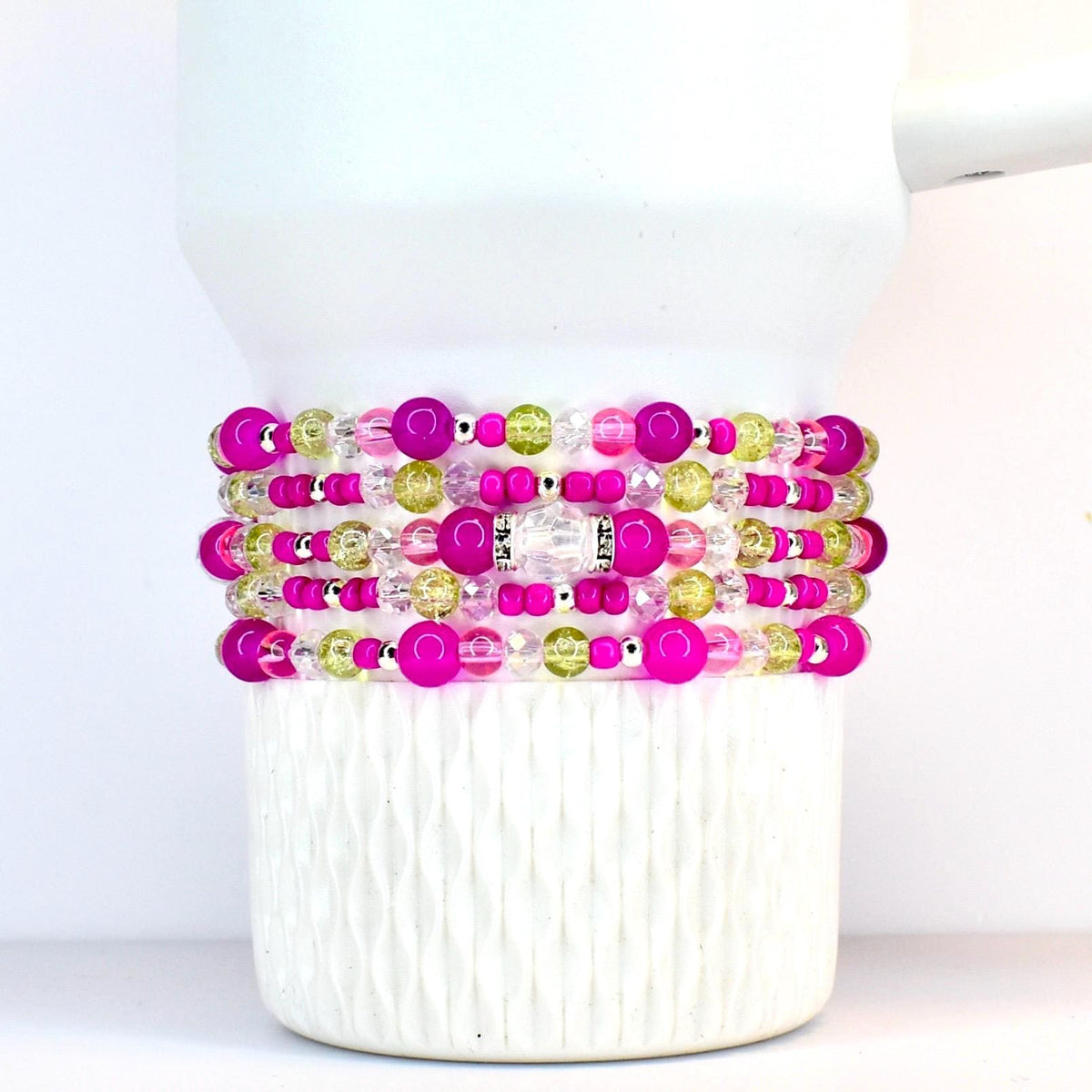 Stack of beaded bracelets in shades of pink, green, and clear. The bracelets are made with various sizes of beads, including large pink orbs, small silver beads, and clear faceted beads. The bracelets are stacked on a white tumbler.