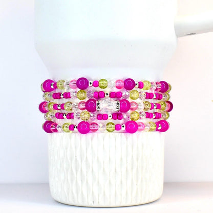 Stack of beaded bracelets in shades of pink, green, and clear. The bracelets are made with various sizes of beads, including large pink orbs, small silver beads, and clear faceted beads. The bracelets are stacked on a white tumbler.