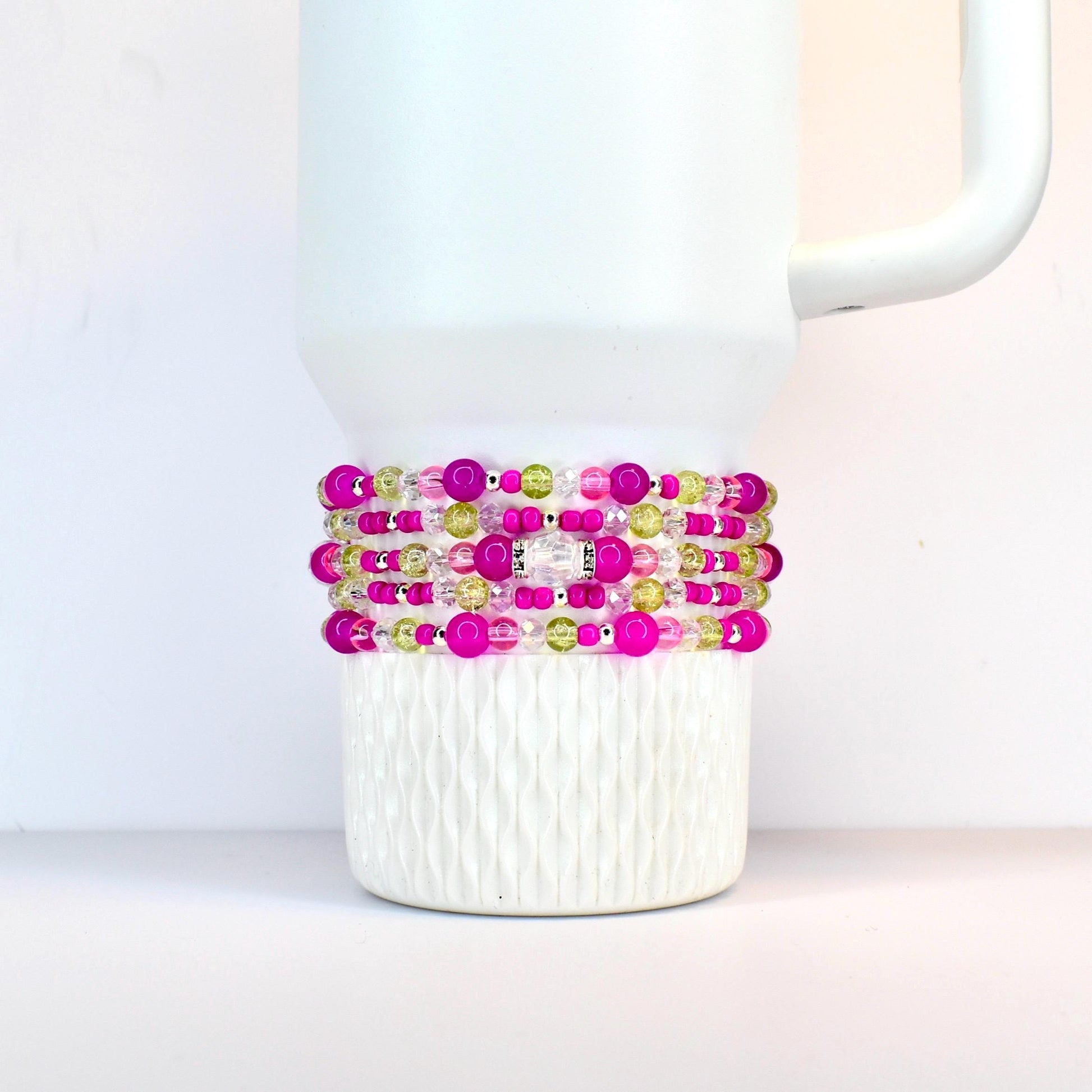A stack of beaded bracelets in shades of pink, green, and clear, wrapped around a white Stanley. The bracelets feature various sizes of beads and a silver-colored accent piece.