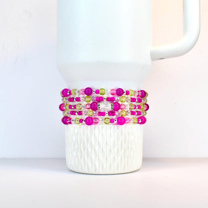 A stack of beaded bracelets in shades of pink, green, and clear, wrapped around a white Stanley. The bracelets feature various sizes of beads and a silver-colored accent piece.