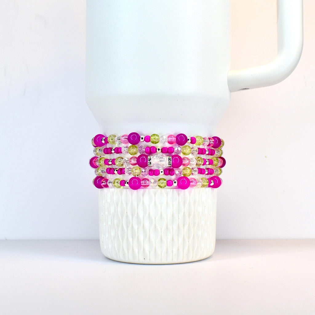 A stack of beaded bracelets in shades of pink, green, and clear, wrapped around a white Stanley. The bracelets feature various sizes of beads and a silver-colored accent piece.
