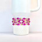 A stack of beaded bracelets in shades of pink, green, and clear, wrapped around a white Stanley. The bracelets feature various sizes of beads and a silver-colored accent piece.