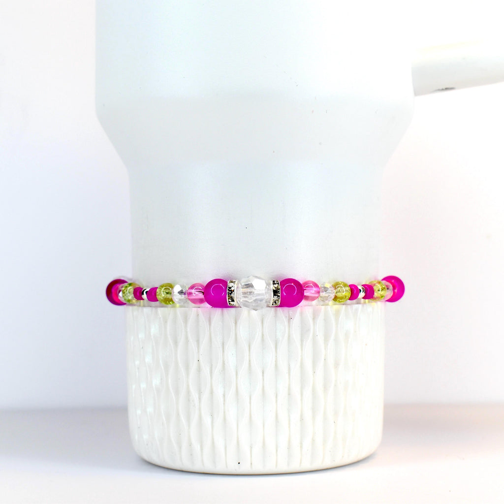 A beaded bracelet featuring a mix of bright pink, clear, and light green beads. The bracelet has a central clear, faceted bead and is displayed on a white tumbler.