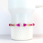 A beaded bracelet featuring a mix of bright pink, clear, and light green beads. The bracelet has a central clear, faceted bead and is displayed on a white tumbler.