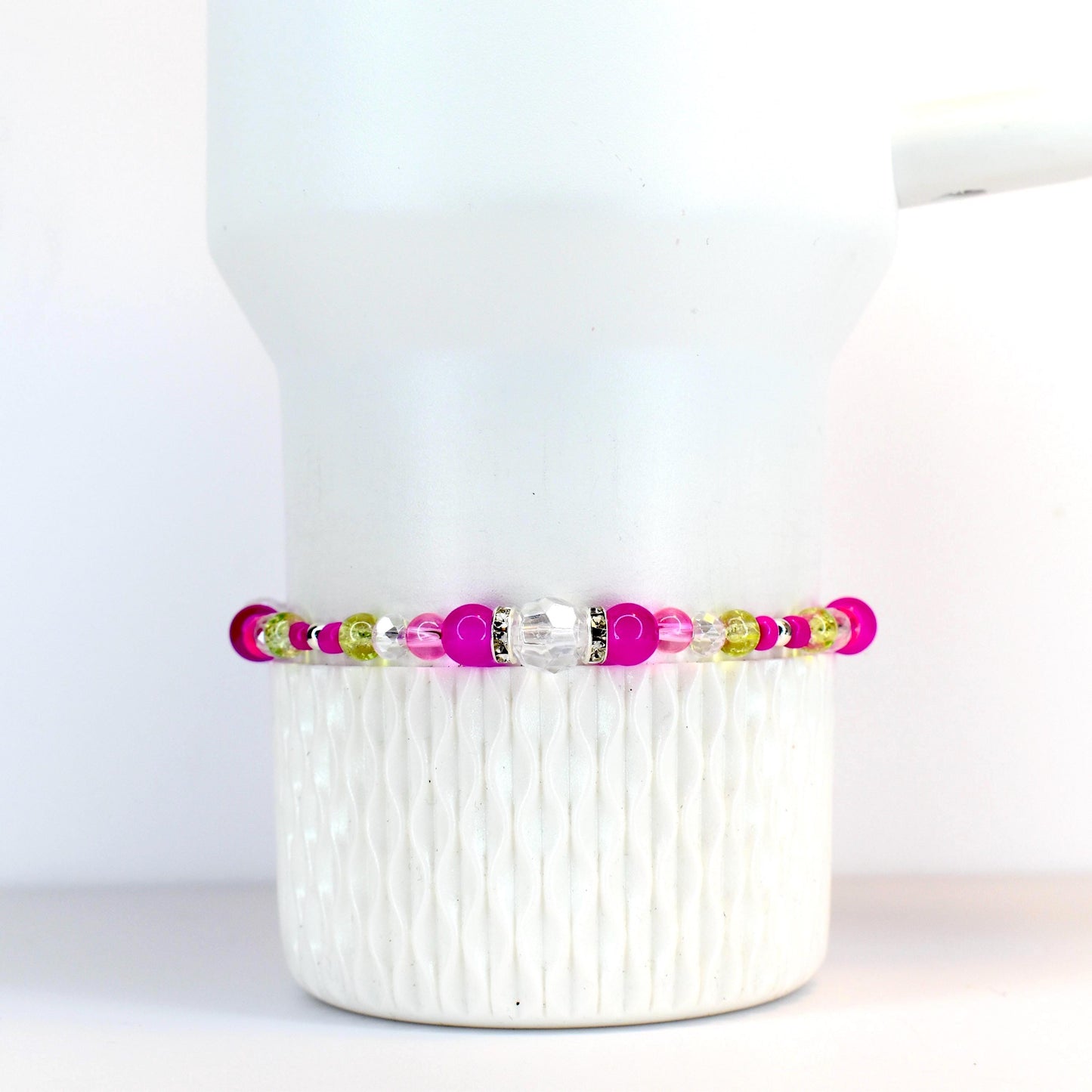 A beaded bracelet featuring a mix of bright pink, clear, and light green beads. The bracelet has a central clear, faceted bead and is displayed on a white tumbler.
