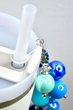 Close-up of a white tumbler with a clear straw and a beaded charm. The charm features a cluster of blue and teal beads in various shades, attached with a silver clasp. The tumbler has a white lid and a silver accent.