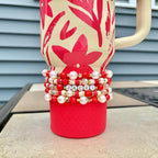 A red and cream-colored tumbler with a floral design and a cream-colored handle. The tumbler is adorned with a stack of beaded bracelets in red, white, and gold, spelling out the name "JACKI".