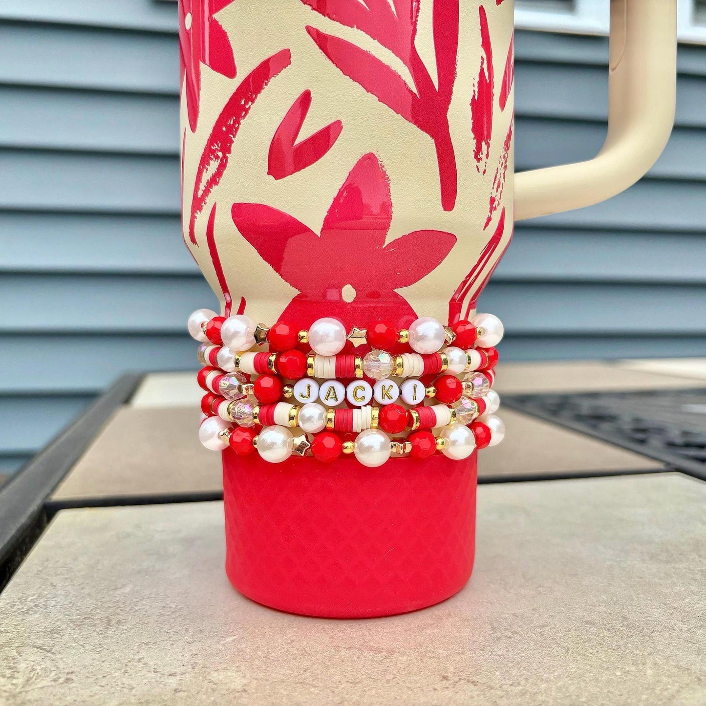 A red and cream-colored tumbler with a floral design and a cream-colored handle. The tumbler is adorned with a stack of beaded bracelets in red, white, and gold, spelling out the name "JACKI".