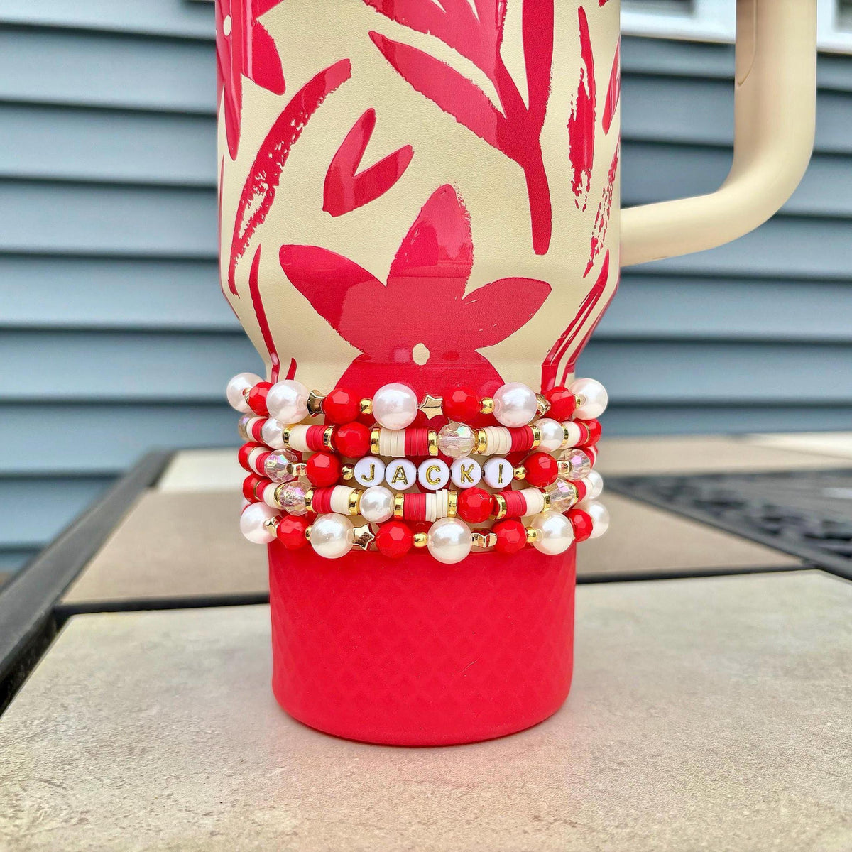 A red and cream-colored tumbler with a floral design and a cream-colored handle. The tumbler is adorned with a stack of beaded bracelets in red, white, and gold, spelling out the name "JACKI".