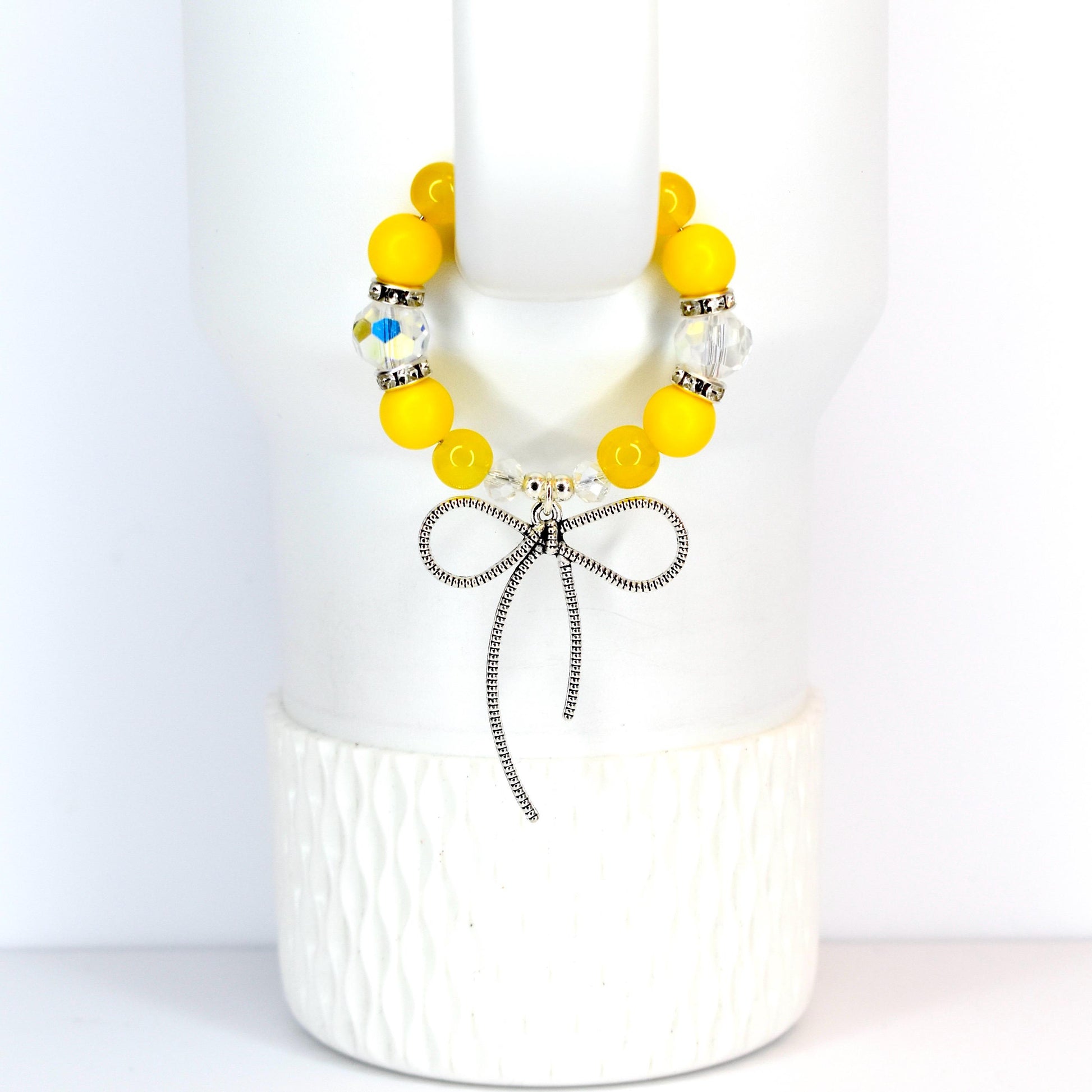 A yellow beaded bracelet with a silver bow charm. The bracelet features large, round yellow beads interspersed with clear, faceted beads. The bow charm is detailed and hangs from the center of the bracelet, creating a delicate and feminine accessory.