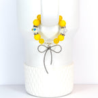 A yellow beaded bracelet with a silver bow charm. The bracelet features large, round yellow beads interspersed with clear, faceted beads. The bow charm is detailed and hangs from the center of the bracelet, creating a delicate and feminine accessory.