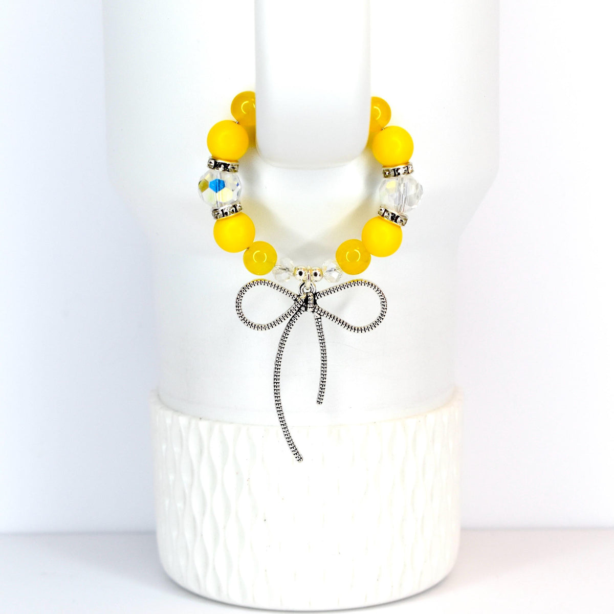 A yellow beaded bracelet with a silver bow charm. The bracelet features large, round yellow beads interspersed with clear, faceted beads. The bow charm is detailed and hangs from the center of the bracelet, creating a delicate and feminine accessory.