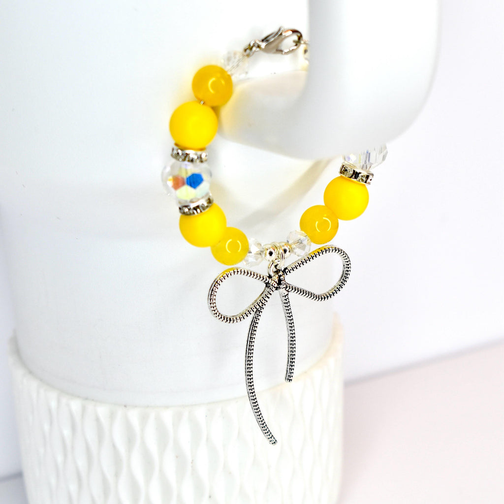 A yellow beaded bracelet with a silver bow charm. The bracelet features bright yellow beads, clear crystal beads, and a decorative bead with colorful accents. The silver bow charm hangs from the center of the bracelet.
