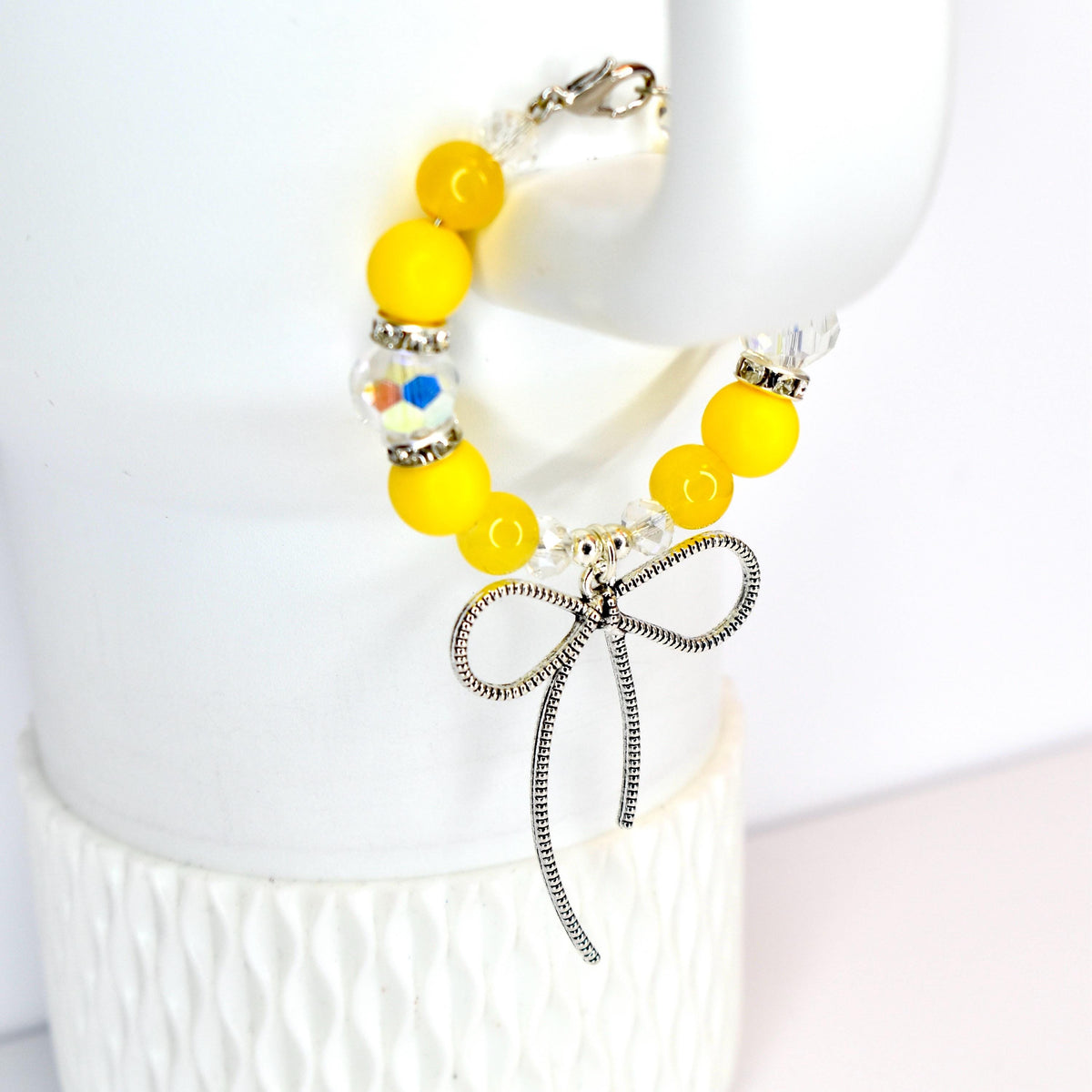 A yellow beaded bracelet with a silver bow charm. The bracelet features bright yellow beads, clear crystal beads, and a decorative bead with colorful accents. The silver bow charm hangs from the center of the bracelet.