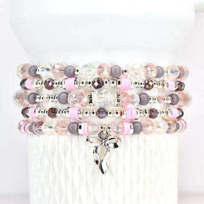 Stack of beaded bracelets in shades of pink, gray, and clear. Each bracelet features a mix of beads, including faceted crystals, round beads, and pink disc beads. A silver bow charm dangles from the bottom bracelet.