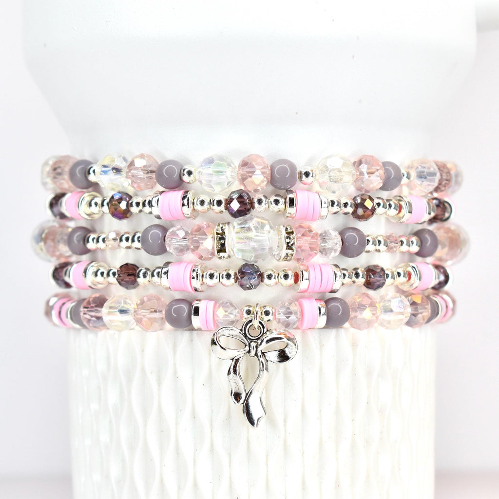 Stack of beaded bracelets in shades of pink, gray, and clear. Each bracelet features a mix of beads, including faceted crystals, round beads, and pink disc beads. A silver bow charm dangles from the bottom bracelet.