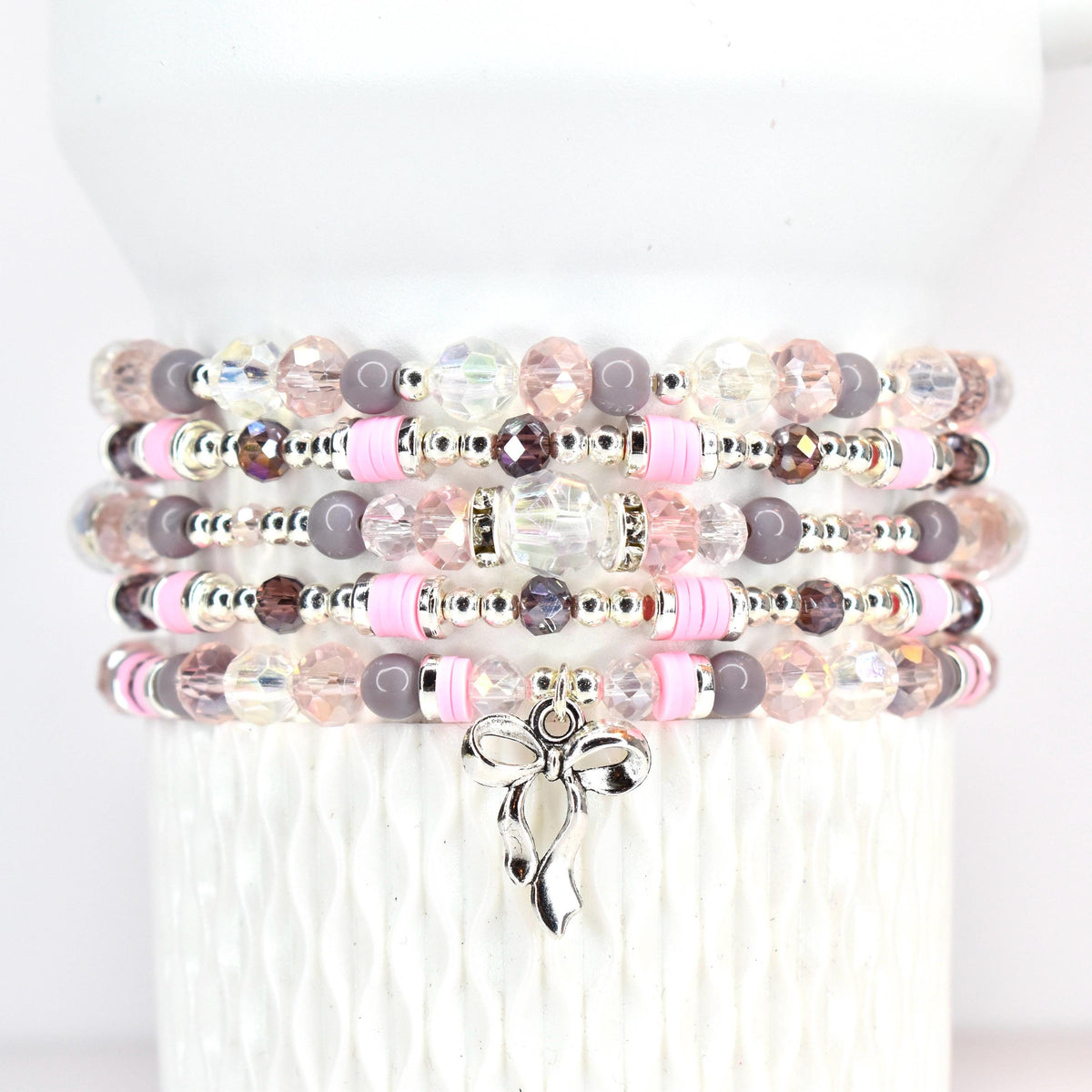 Stack of beaded bracelets in shades of pink, gray, and clear. Each bracelet features a mix of beads, including faceted crystals, round beads, and pink disc beads. A silver bow charm dangles from the bottom bracelet.