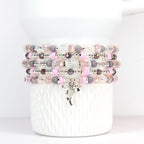 Stack of beaded bracelets in shades of pink, plum purple, and clear. Each bracelet features a mix of beads, including round, faceted, and spacer beads. A silver-tone bow charm hangs from the bottom bracelet. The bracelets are displayed on a white ceramic container.