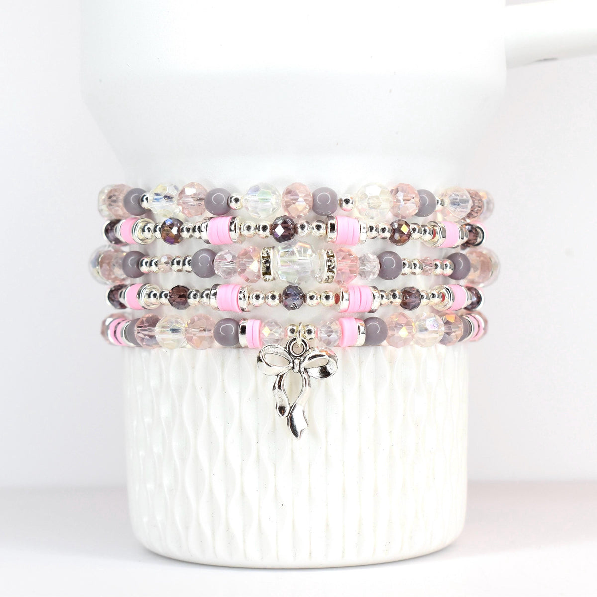 Stack of beaded bracelets in shades of pink, plum purple, and clear. Each bracelet features a mix of beads, including round, faceted, and spacer beads. A silver-tone bow charm hangs from the bottom bracelet. The bracelets are displayed on a white ceramic container.